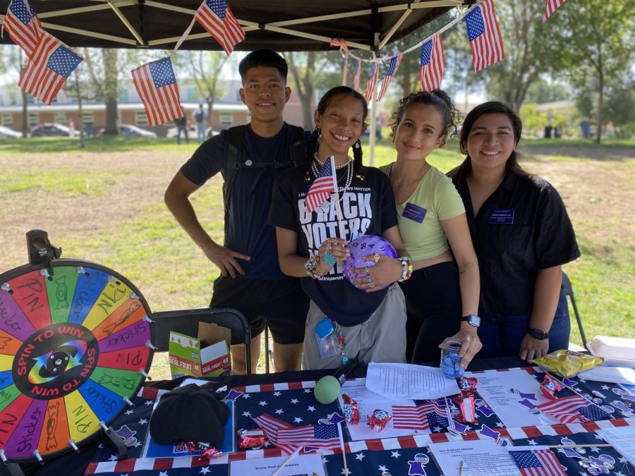 Four people stand behind a table decorated with American flag pins, vote stickers, and a spin to win wheel. The table also has QR codes with voting information.